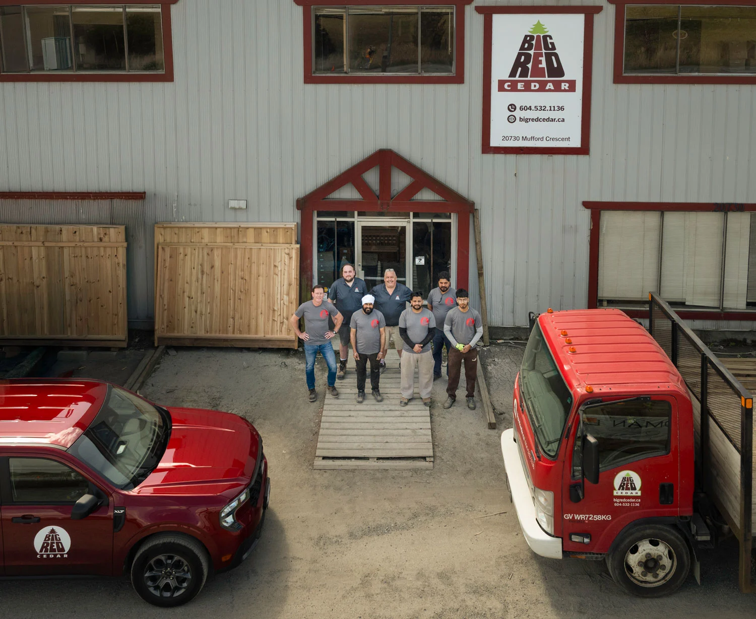 Team of workers in gray shirts stand outside a workshop with red trim and a Big Red Cedar sign on the building. Behind them are wooden crates and red trucks nearby.