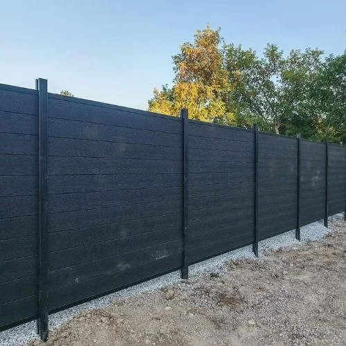 Long black privacy fence with evenly spaced posts along a gravel base, trees in the background at the top edge of the image.