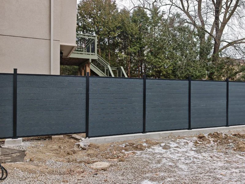Blue-gray privacy fence panels with black posts along a construction area beside a beige building and bare ground.