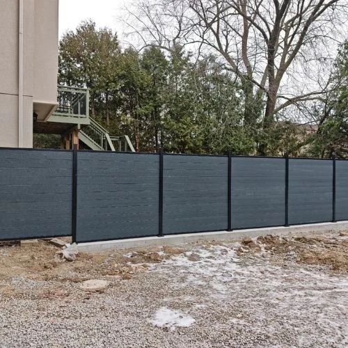 Blue privacy fence panels run along a construction area beside a building, with bare trees behind and snow patches on the ground.