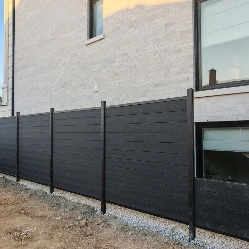 Dark gray privacy fence along a modern light brick building, with vertical posts and gravel base.
