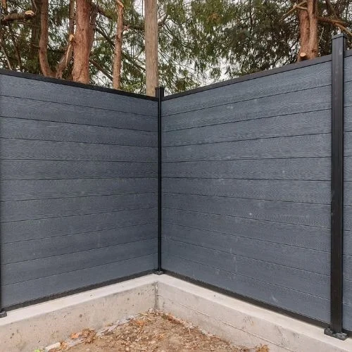 Corner section of dark gray privacy fence panels meeting at a right angle, supported by black posts, with a concrete base and trees visible behind.