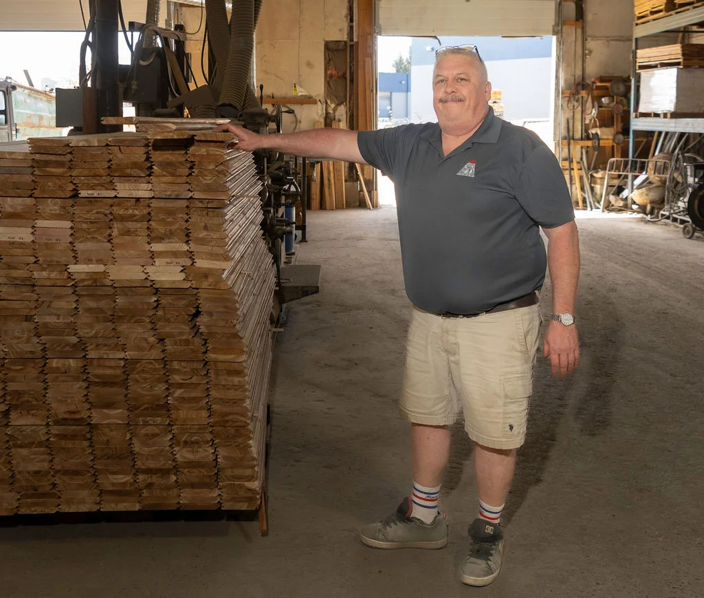 Man in a dark polo shirt leaning on a tall stack of plywood sheets in a workshop bay.