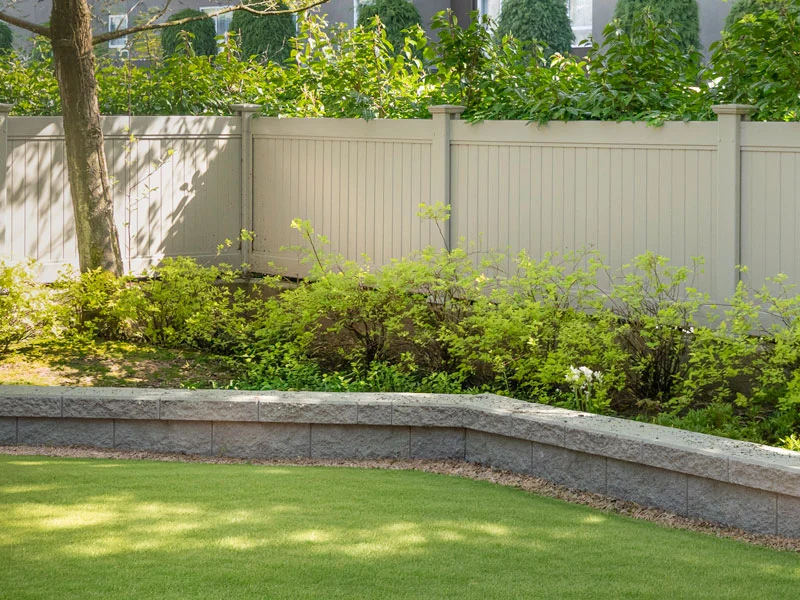 Backyard landscaped with a low gray stone raised planter border, green shrubs, and a white privacy fence behind a well-kept lawn.