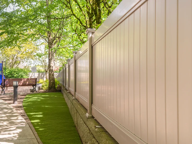 Beige fence along a sidewalk in a sunny park with trees, grass, and a bench nearby