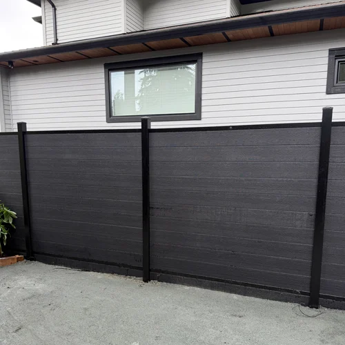 Black privacy fence panels run along a gray-siding house, with a rectangular window visible above the fence.