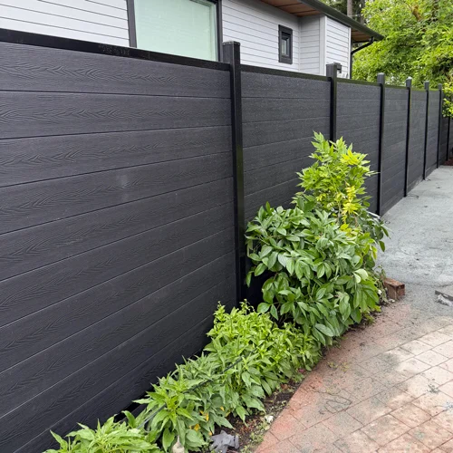 Long black privacy fence with evenly spaced posts along a walkway, with green shrubs at its base on the side yard.