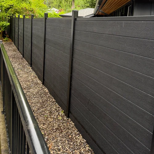 Long row of dark gray privacy fence panels with evenly spaced vertical posts, running along a gravel path beside a house and green trees in the background.